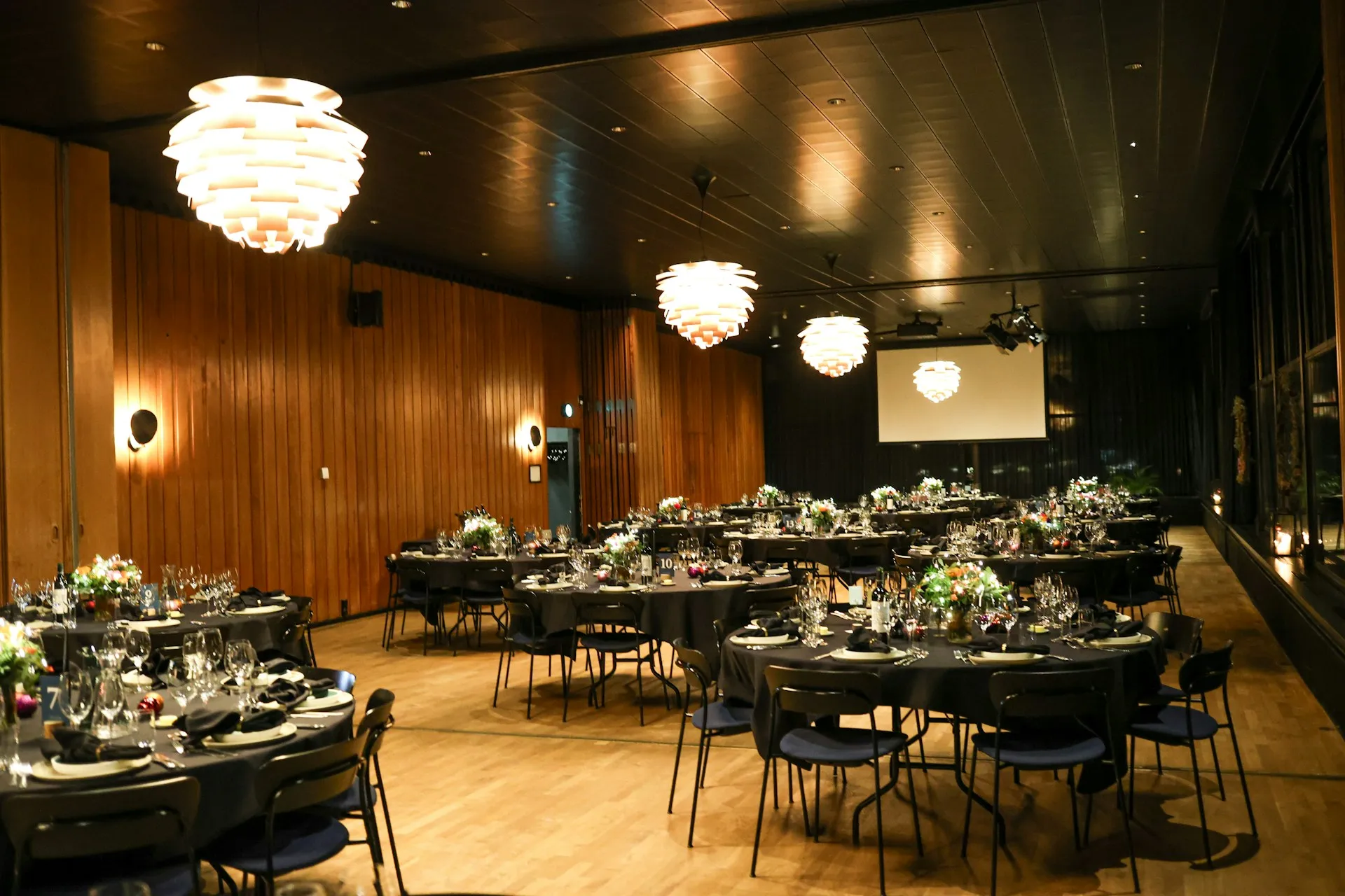 Sala allestita per una cena di gala aziendale, con tavoli rotondi apparecchiati e schermo per le proiezioni